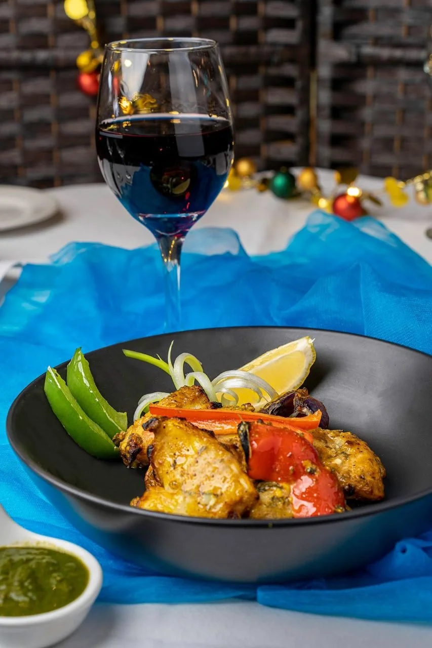 A table setting with a glass of red wine, a black bowl of grilled chicken with vegetables and lemon wedge, and a small bowl of green sauce, with holiday decorations in the background.