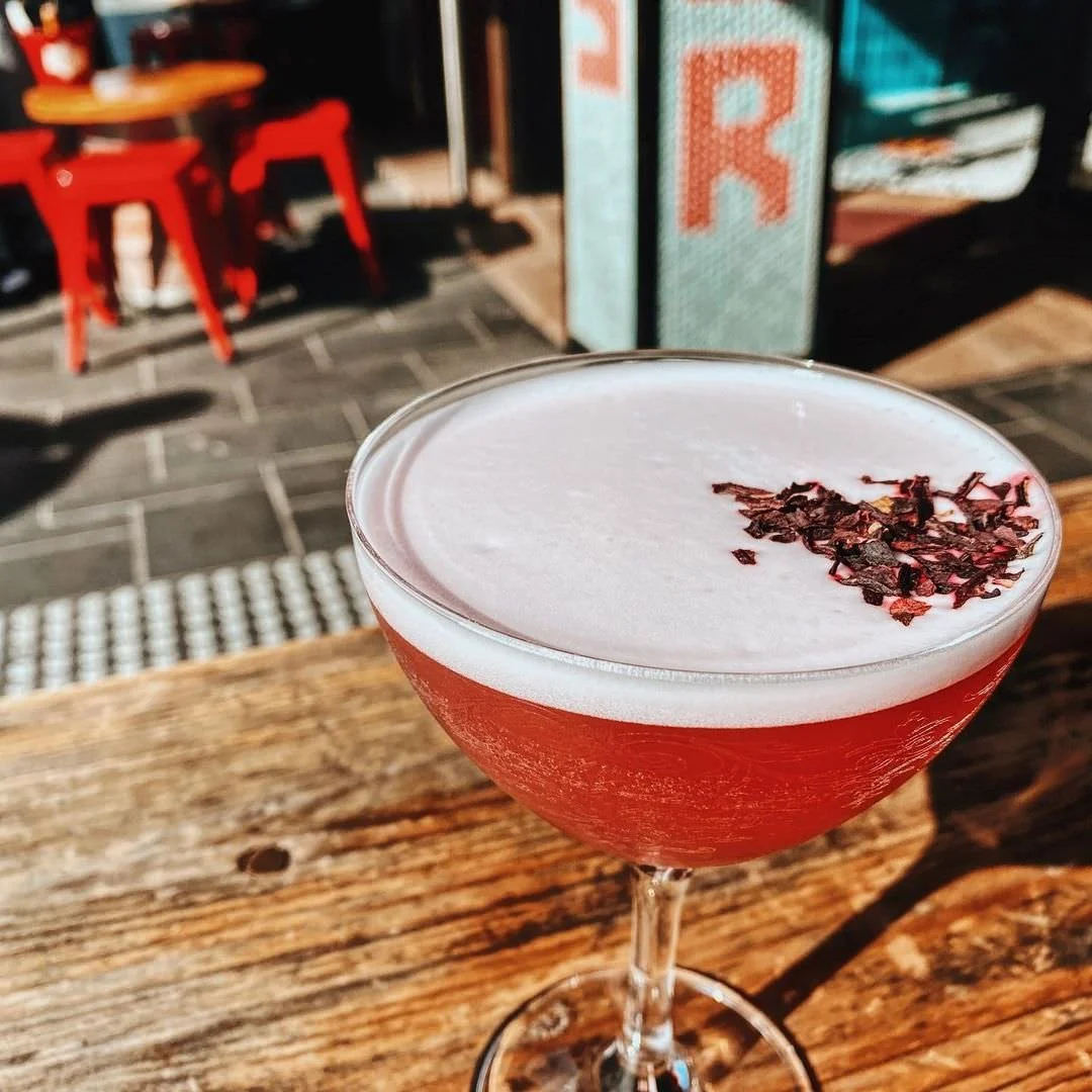 A pink cocktail with a foamy white top and dark red flower petals garnish, served in a coupe glass on a wooden table outdoors.