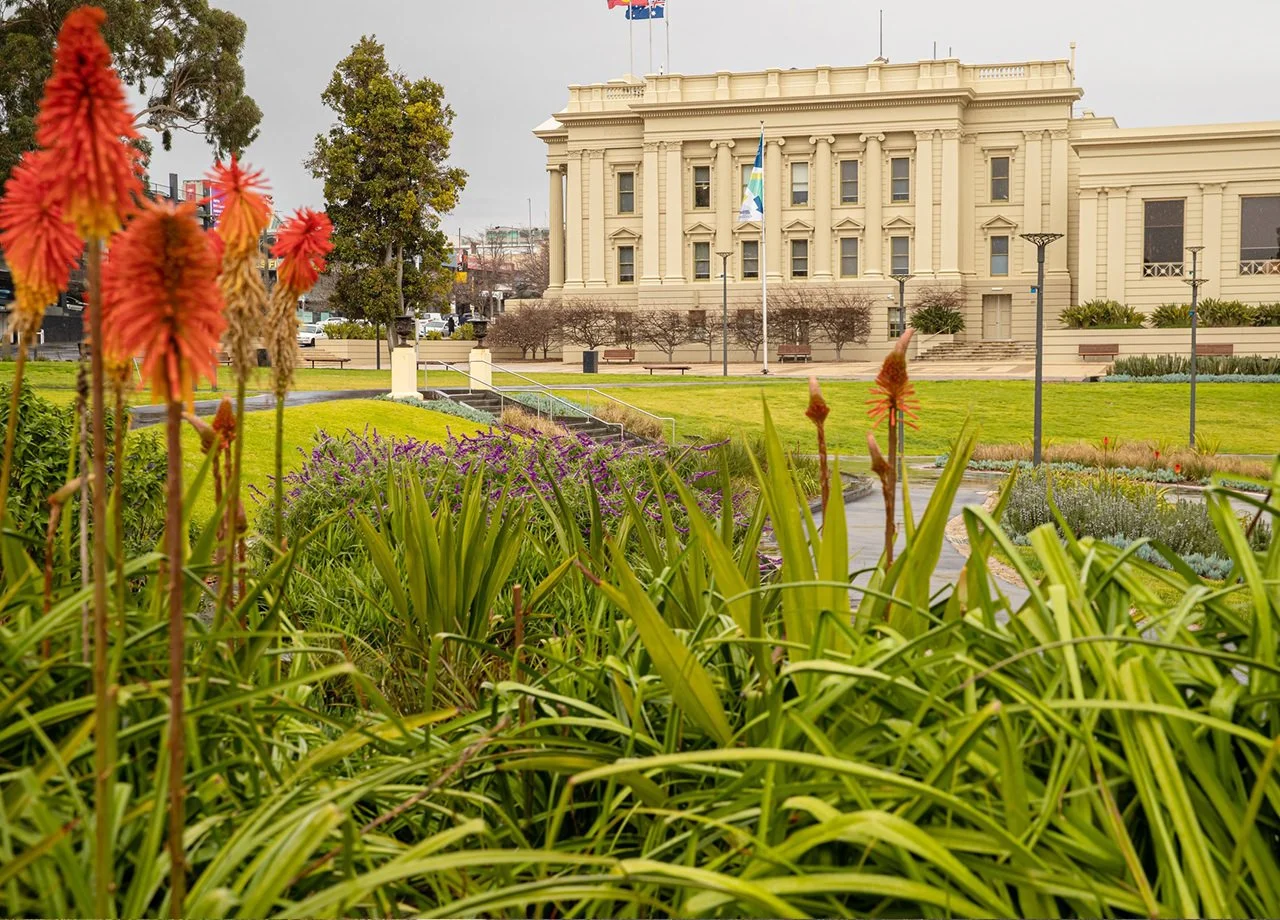 A government or historic building with a cream-colored facade, surrounded by a well-maintained garden with flowers and greenery in the foreground, and cloudy sky overhead.