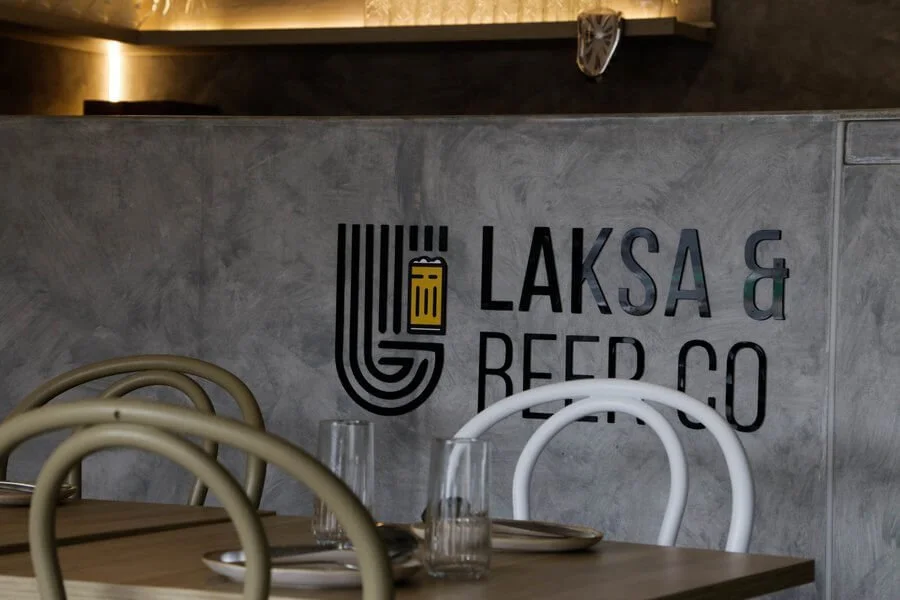 Decorative wall with the sign "Laks & Reed Co" and a stylized logo, in a restaurant with tables set with glasses and plates.