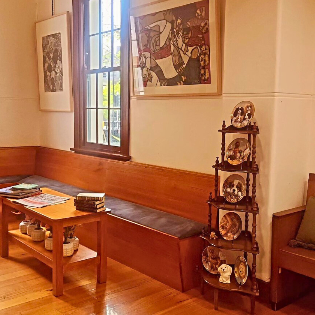 Cozy corner of a room with wooden bench seating, a wooden coffee table with books and magazines, a window with sunlight, and a wooden shelf displaying decorative plates with dog images.