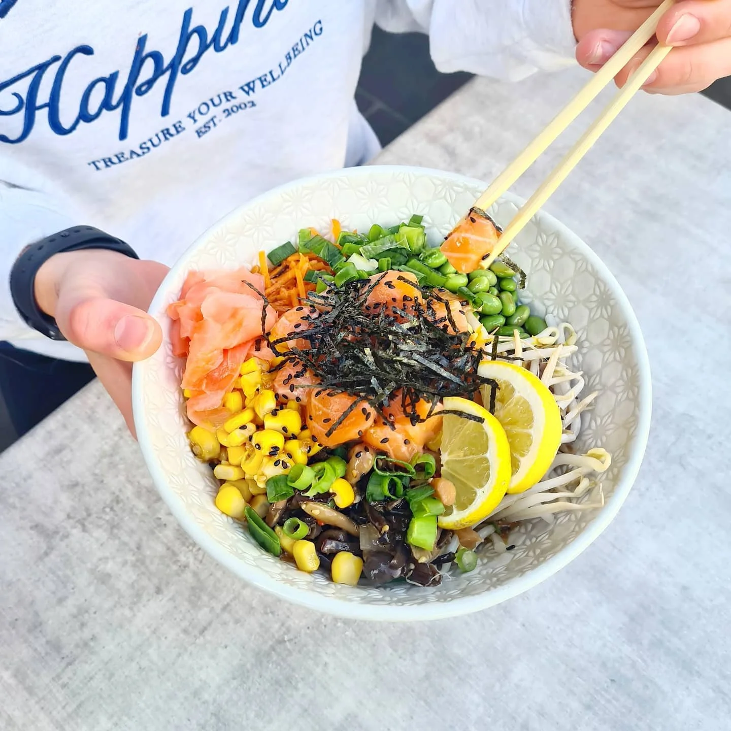 Person holding a bowl of poke with salmon, lemon slices, green onions, edamame, shredded carrots, seaweed, and chopped green vegetables, using chopsticks.