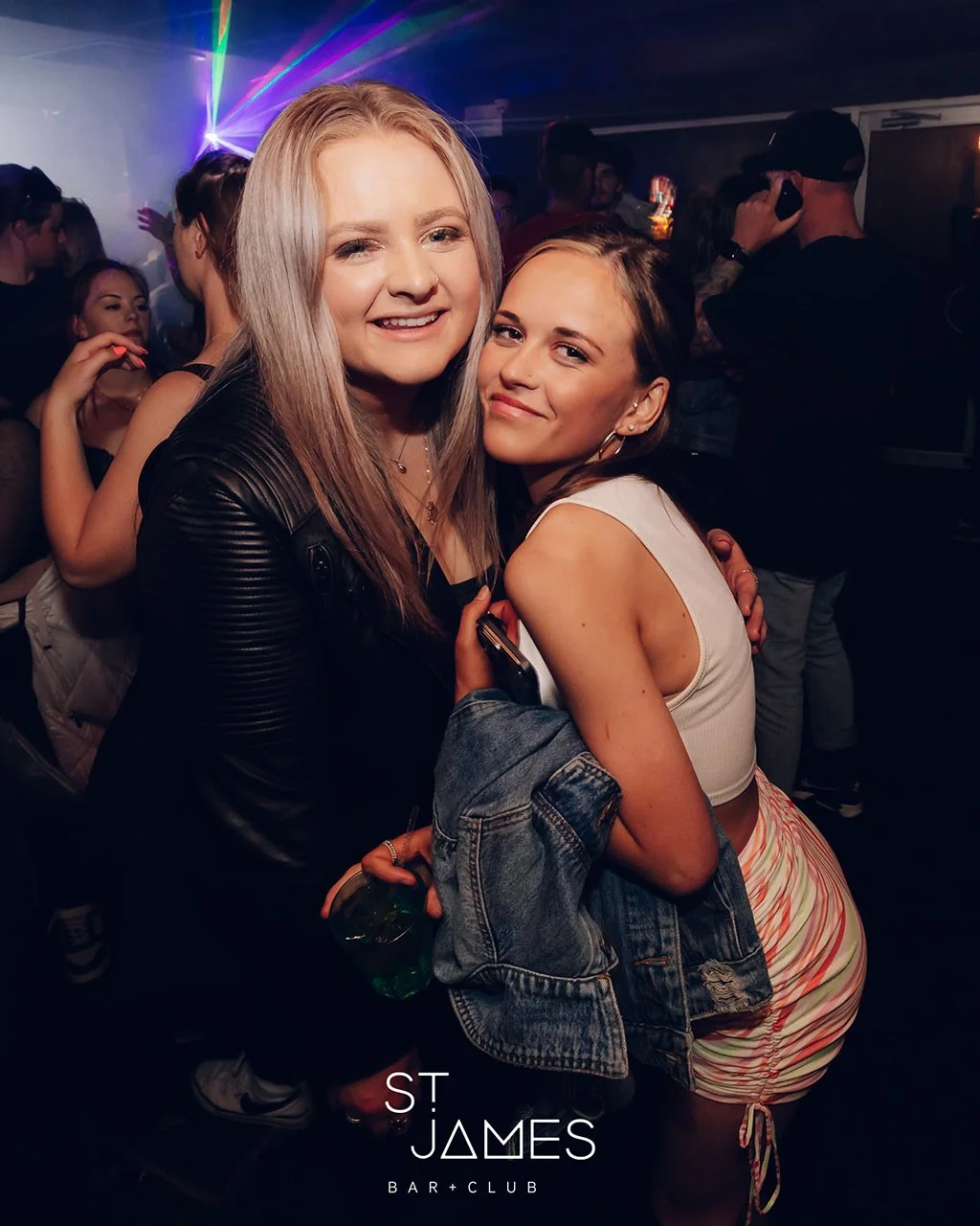 Two women smiling and hugging at a nightclub, with colorful lights and other partygoers in the background.