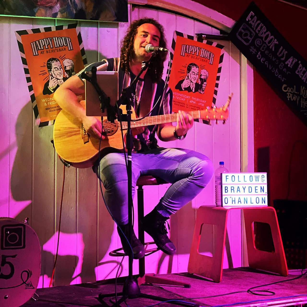 A man with curly hair, dressed in a striped shirt and light jeans, playing an acoustic guitar and singing into a microphone on a small stage in a bar. Posters advertising happy hour are on the wall behind him, and a sign on a stool encourages followi