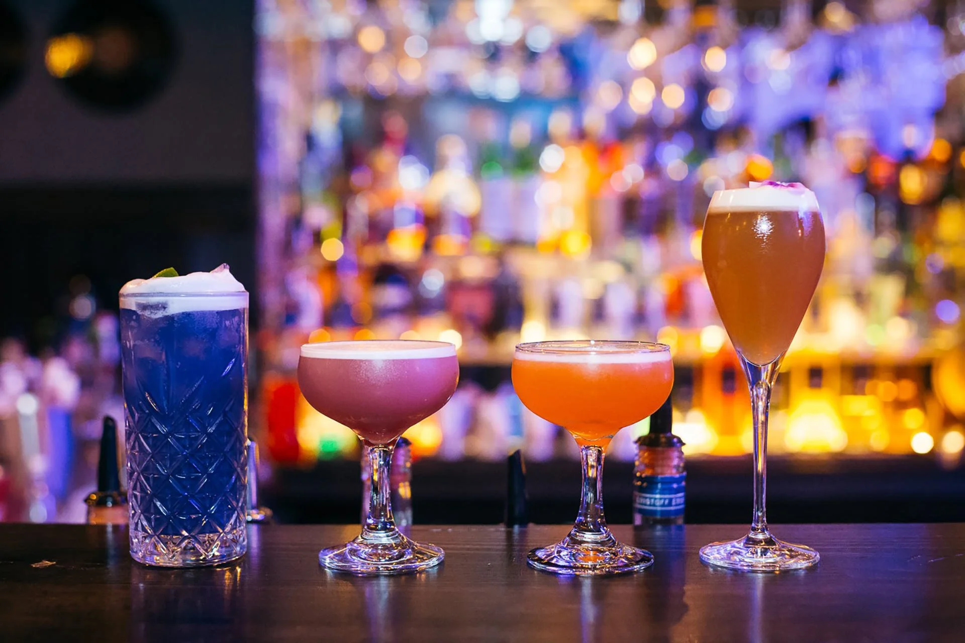Four colorful cocktails on a bar counter with a blurred, vibrant bar backdrop.