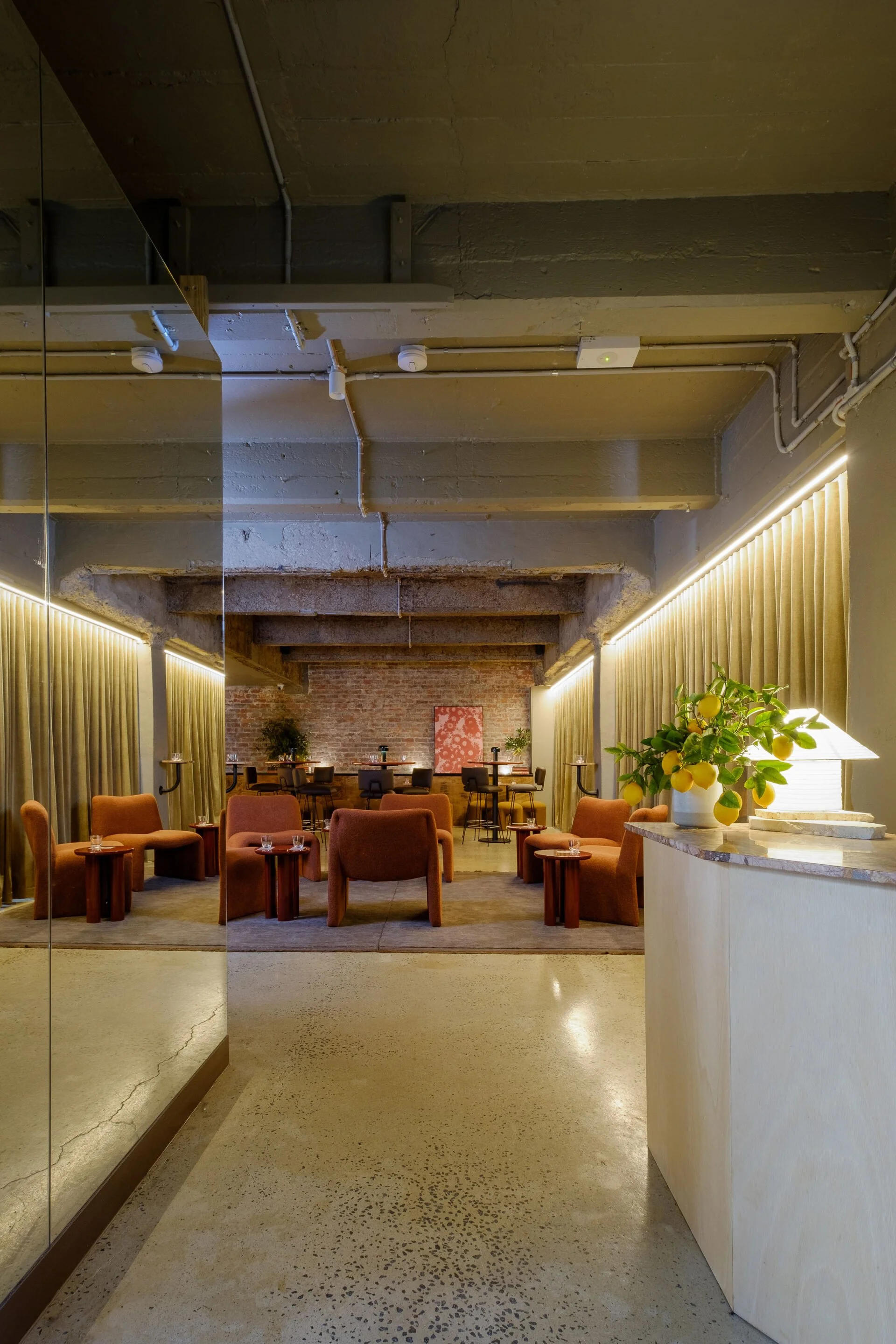 Interior of a cozy lounge area with brown armchairs and small side tables, decorated with yellow lemons in a vase, illuminated by warm lighting, with exposed brick wall and curtains.