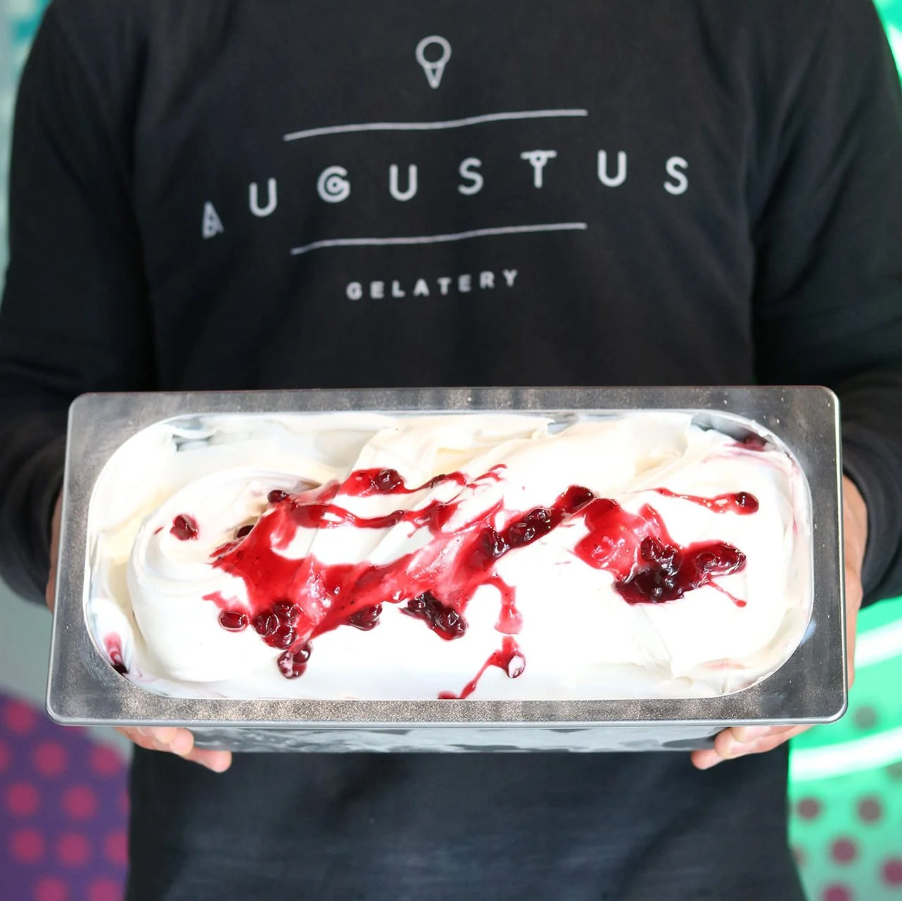 Person holding a rectangular tray of white gelato topped with red berry sauce and whole berries, in front of a person wearing a black shirt with 'Augustus Gelatery' written on it.
