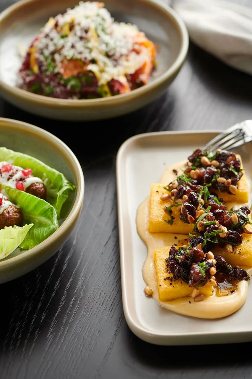 Three dishes on a dark wooden table: a bowl of salad with green lettuce, meatballs, and red pomegranate seeds; a bowl of a grain salad with vegetables; a plate of polenta with a vegetable topping and pine nuts.