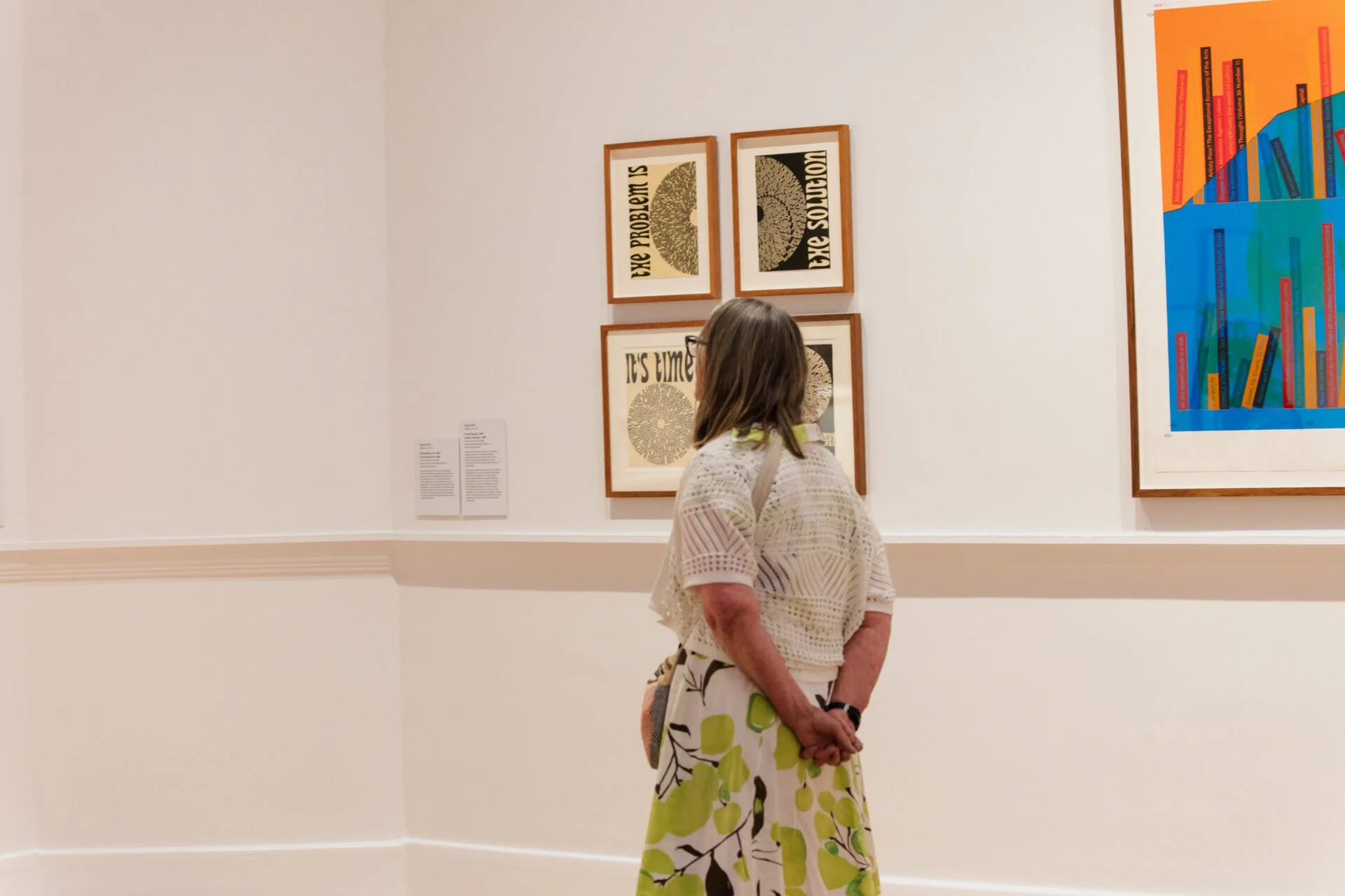 Woman with shoulder-length hair, glasses, wearing a white lace top and a yellow skirt with green leaves, standing in an art gallery, observing framed artwork on the wall.