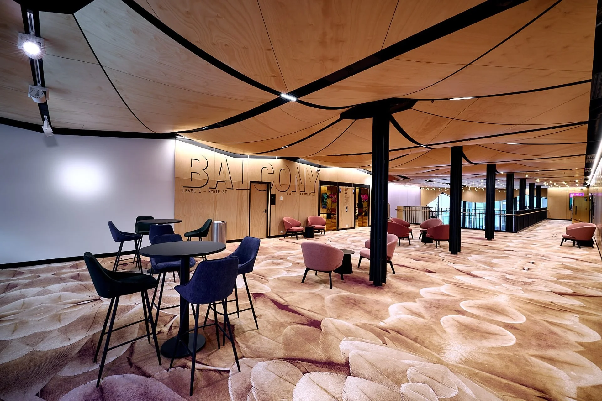 Modern interior lobby area with pink and dark blue chairs, standing tables, wooden ceiling with black structural elements, and a patterned carpet. Sign reads "BALTICON."