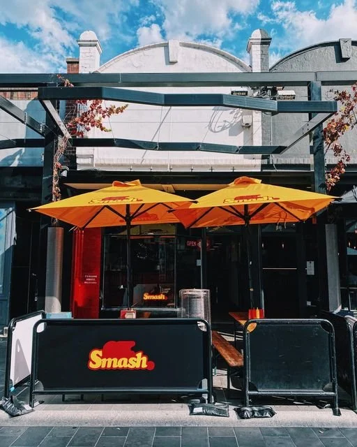Outdoor seating area of a restaurant with two yellow umbrellas, a black barrier with the restaurant name "Smash," and a glass entrance door, under a blue sky with clouds.