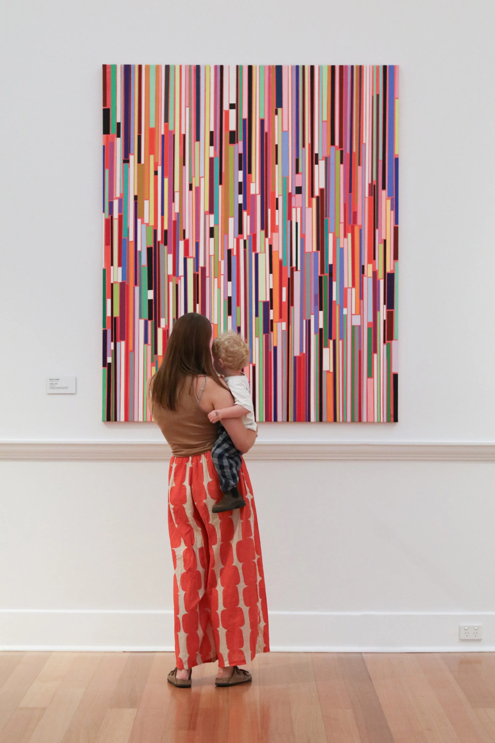A woman holding a child and looking at an abstract colorful painting in a gallery.