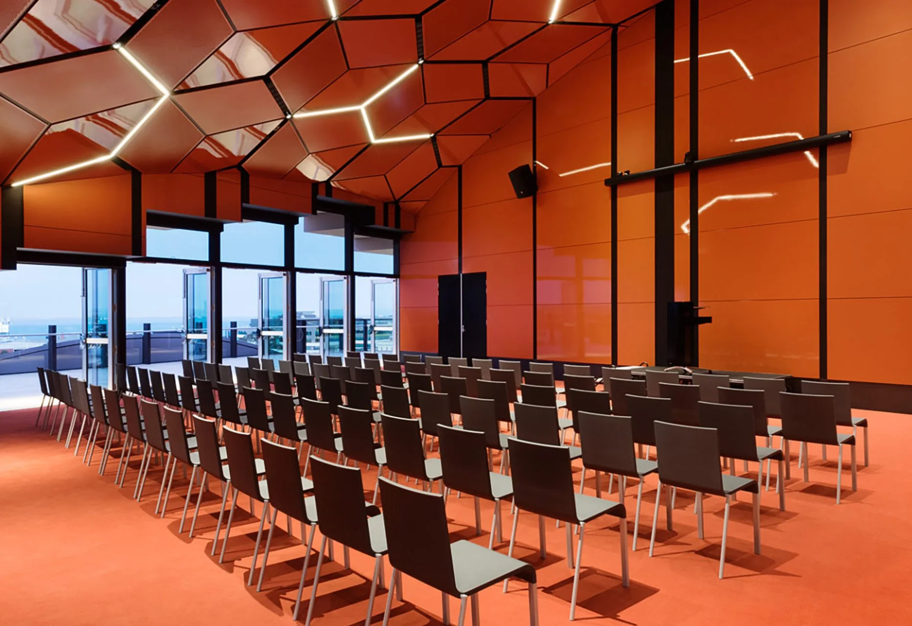 Modern conference room with orange walls, black chairs arranged in rows, and a ceiling with geometric lighting, overlooking a balcony with city and ocean views.