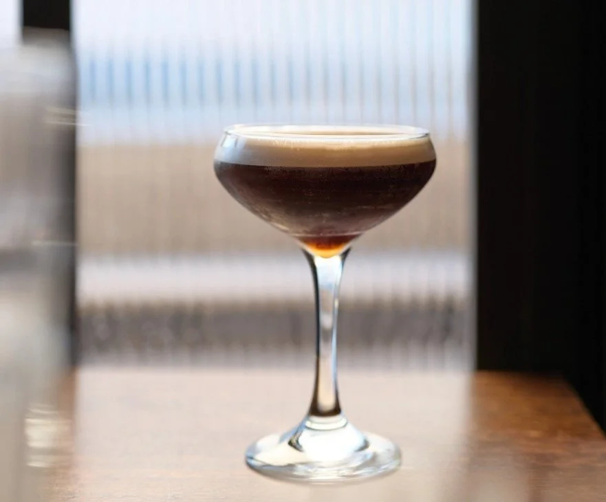 A cocktail glass with a dark-colored drink and a frothy top on a wooden surface, with a blurred window in the background.