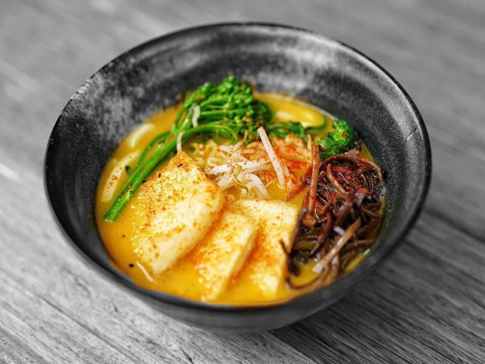 A bowl of ramen soup with tofu, green vegetables, and seaweed on a gray wooden surface.