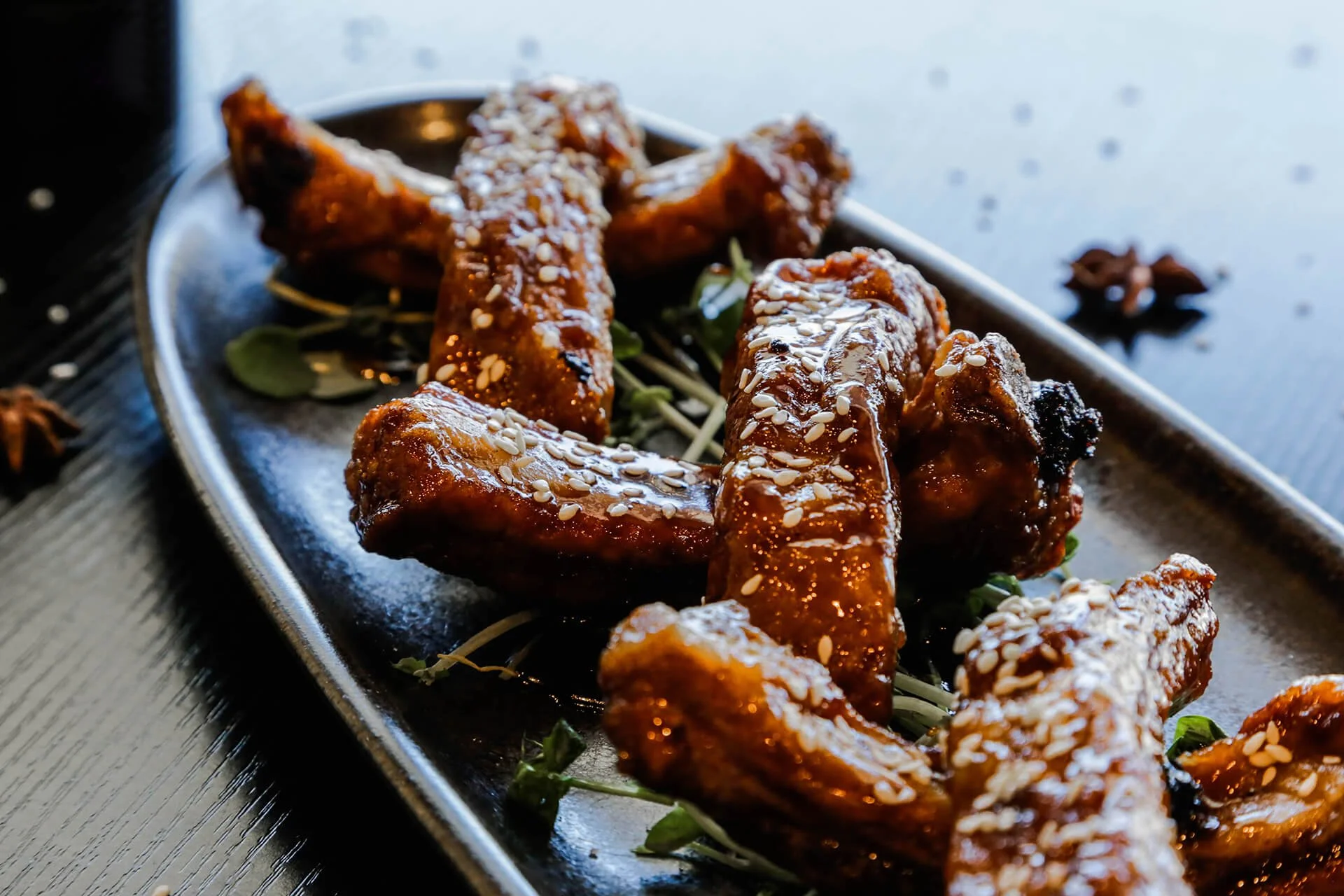 Close-up of glazed chicken wings sprinkled with sesame seeds served on a black rectangular plate.