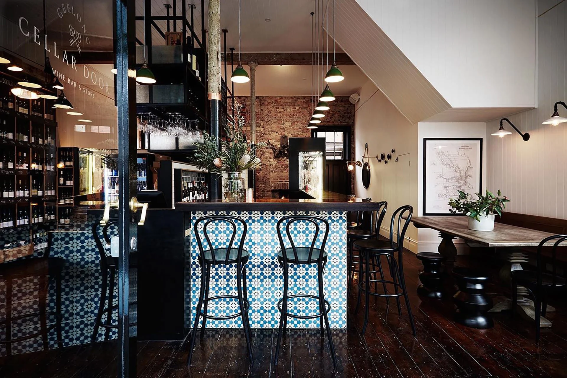 Interior of a cozy wine bar with a patterned blue and white tiled counter, black barstools, a wooden table with a plant, wine bottles on shelves, and soft lighting.