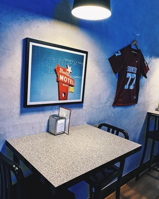 Indoor seating area with a speckled table, three black chairs, a framed vintage neon motel sign, a sports jersey hanging on the wall, and a ceiling light casting light on the wall.