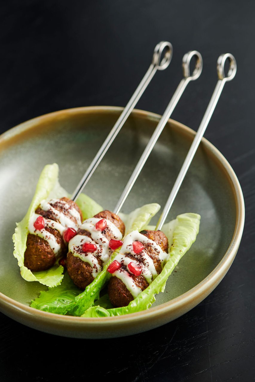 Three skewers of meatballs drizzled with white sauce and garnished with pomegranate seeds, served on lettuce leaves in a ceramic bowl.