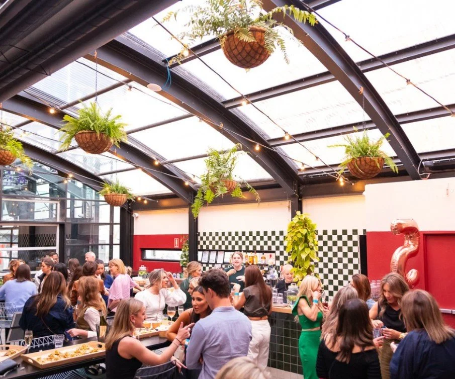 Indoor bar with hanging planters, string lights, and a checkered wall, crowded with people socializing and enjoying drinks.