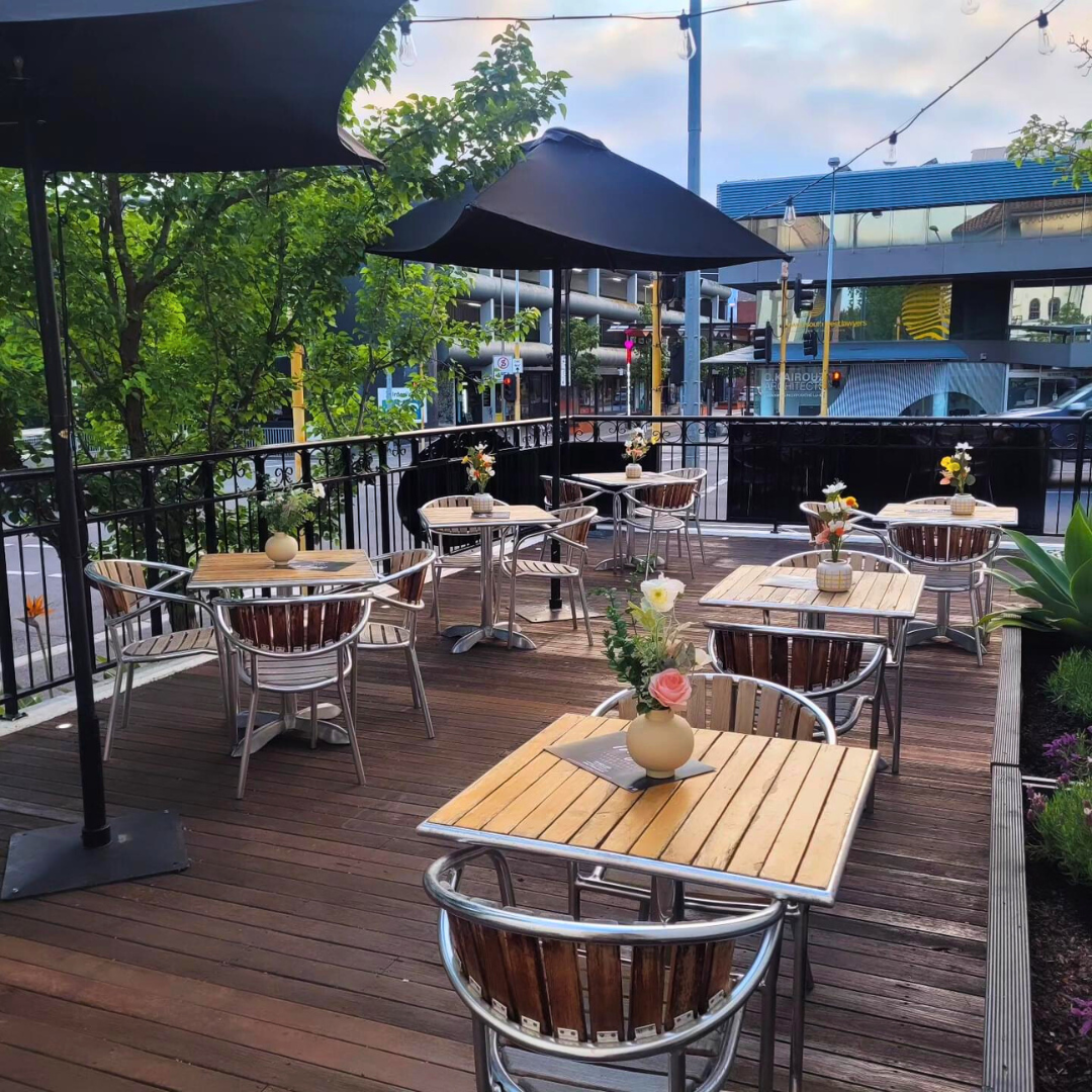 Outdoor patio with wooden tables, metal chairs, and vases of flowers, shaded by large umbrellas, set in an urban area with greenery and city buildings visible in the background.