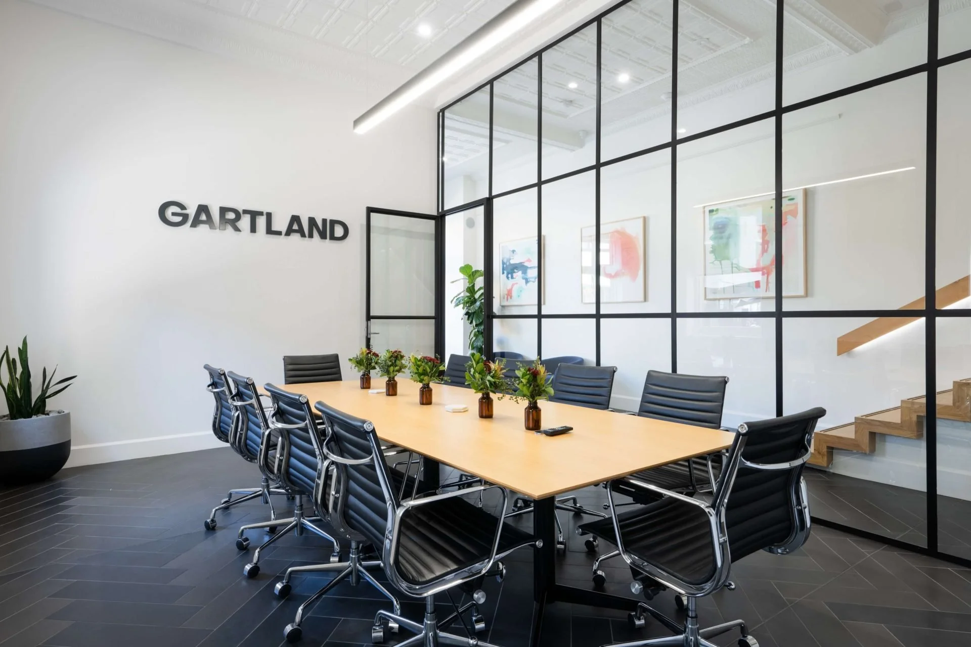 Modern conference room with a large wooden table, black leather chairs, and decorative plants, featuring a glass-walled partition and the word 'GARTLAND' on the white wall.
