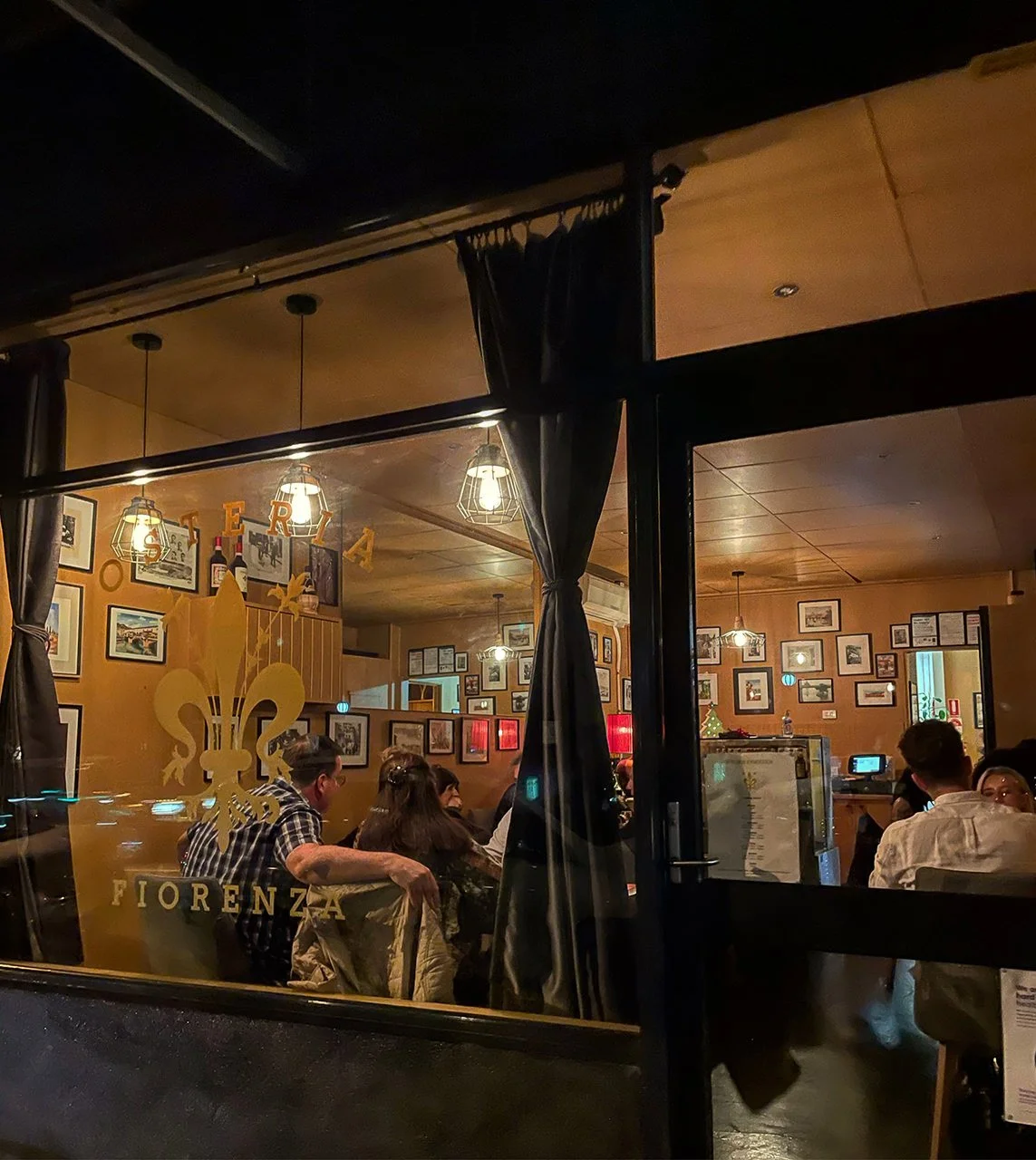 View of a cozy restaurant interior through a glass window with a gold fleur-de-lis logo, warm lighting, framed pictures on the walls, and patrons dining inside.