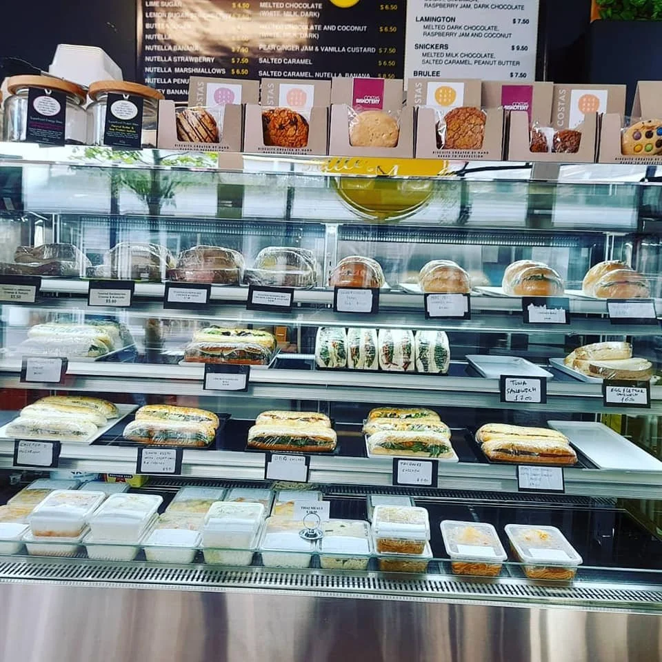 Display case filled with various sandwiches, baked goods, and pastries, with a menu board in the background at a cafe or bakery.