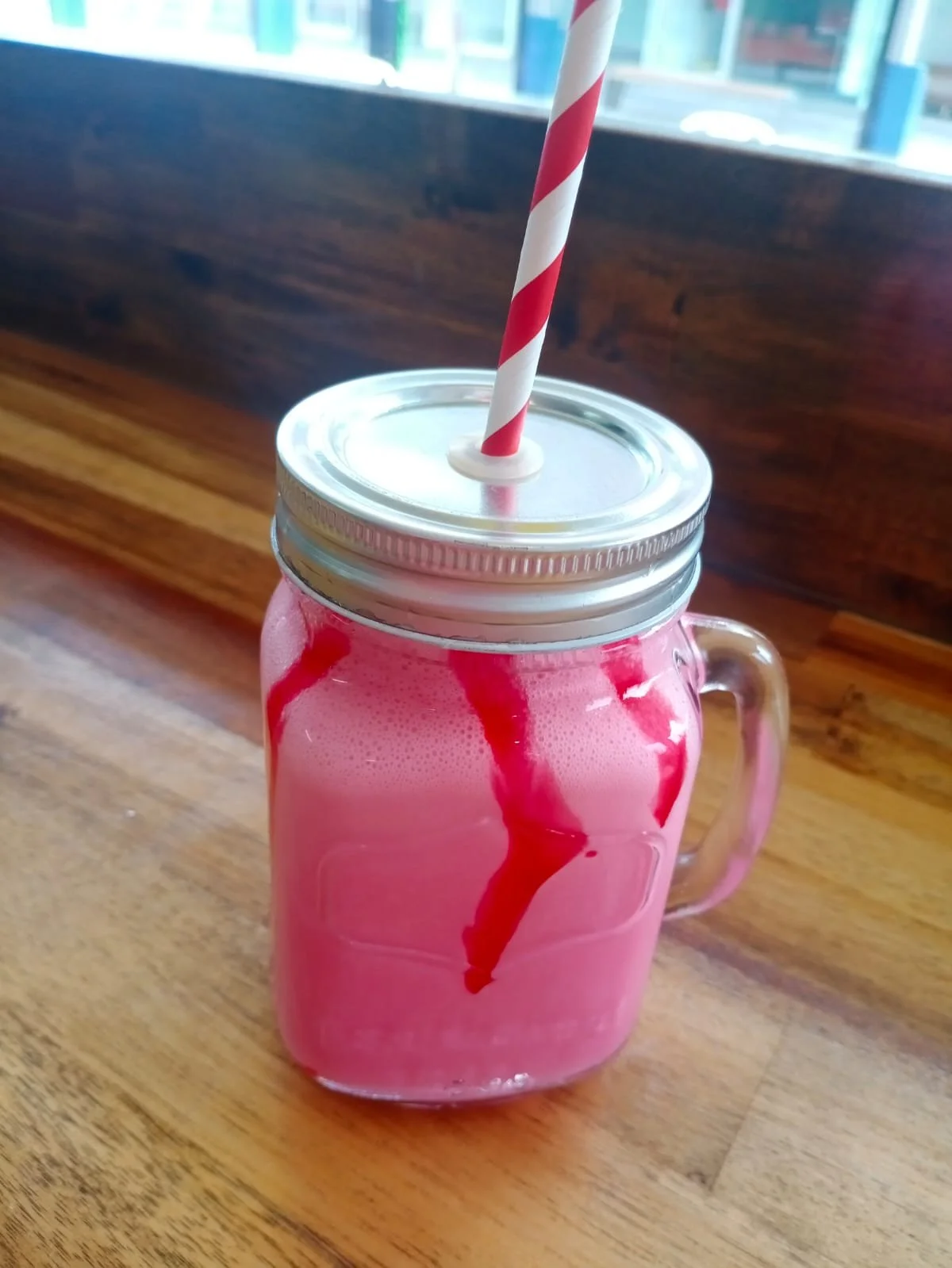 Pink beverage in a mason jar with a metal lid and a red and white striped straw, on a wooden table near a window.