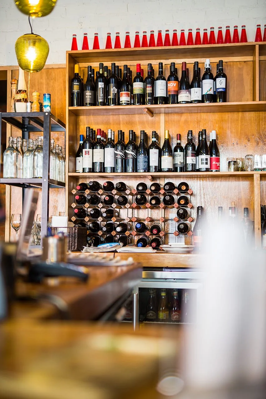 Wooden shelves stocked with various bottles of wine and champagne, with wine glasses and shot glasses nearby, in a bar or restaurant setting.