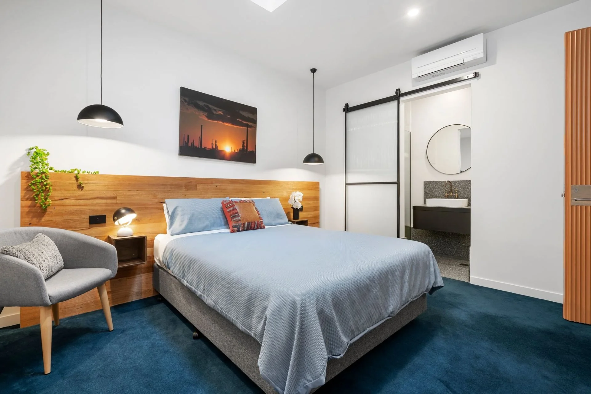 Modern hotel room with a gray bed, wooden headboard, black lamps, gray armchair, and a view of a bathroom sink with mirror, and a sliding door leading to a bathroom