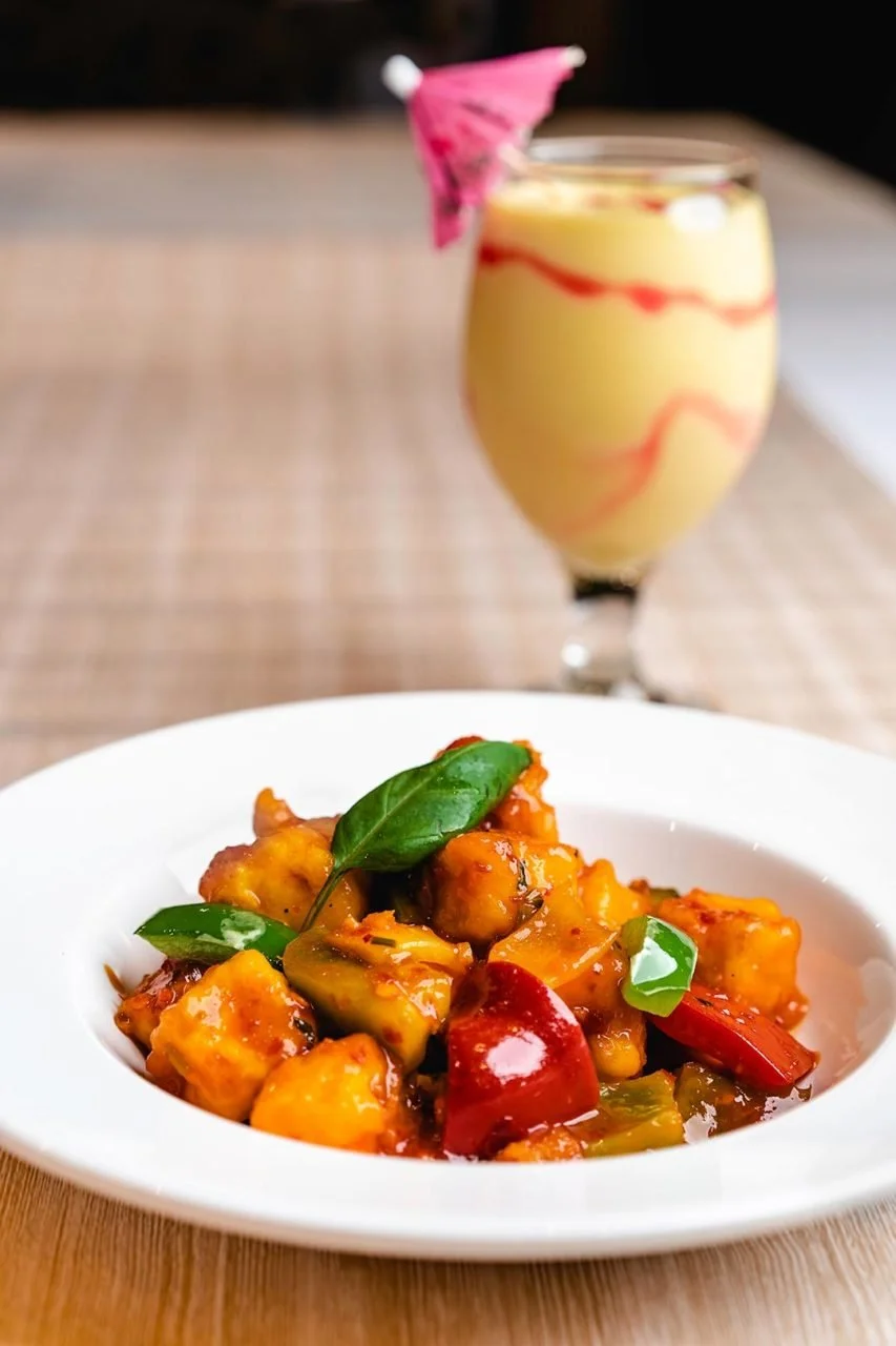 A white bowl of sweet and sour chicken with green basil garnish, and a pink cocktail with a small umbrella in a glass in the background.