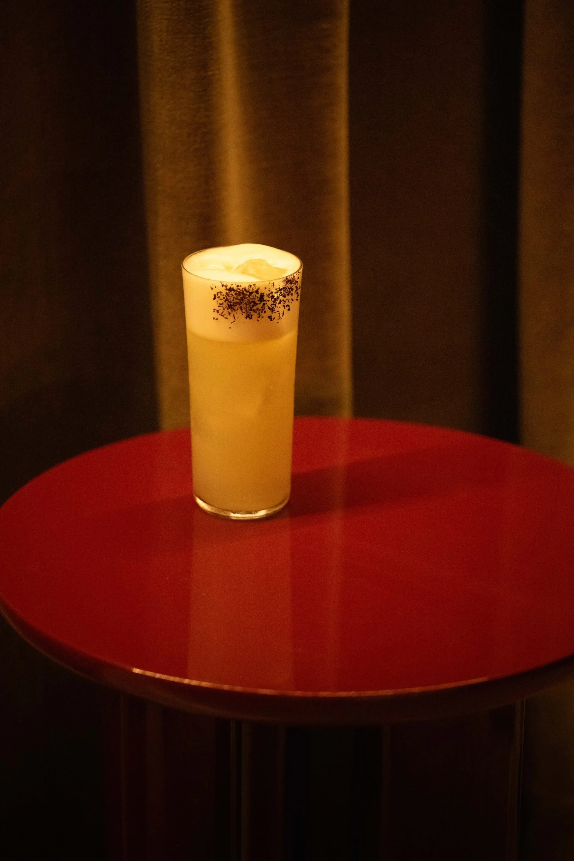 A tall glass of yellow cocktail with a foamy top, garnished with black sprinkles, on a red table against dark curtains.