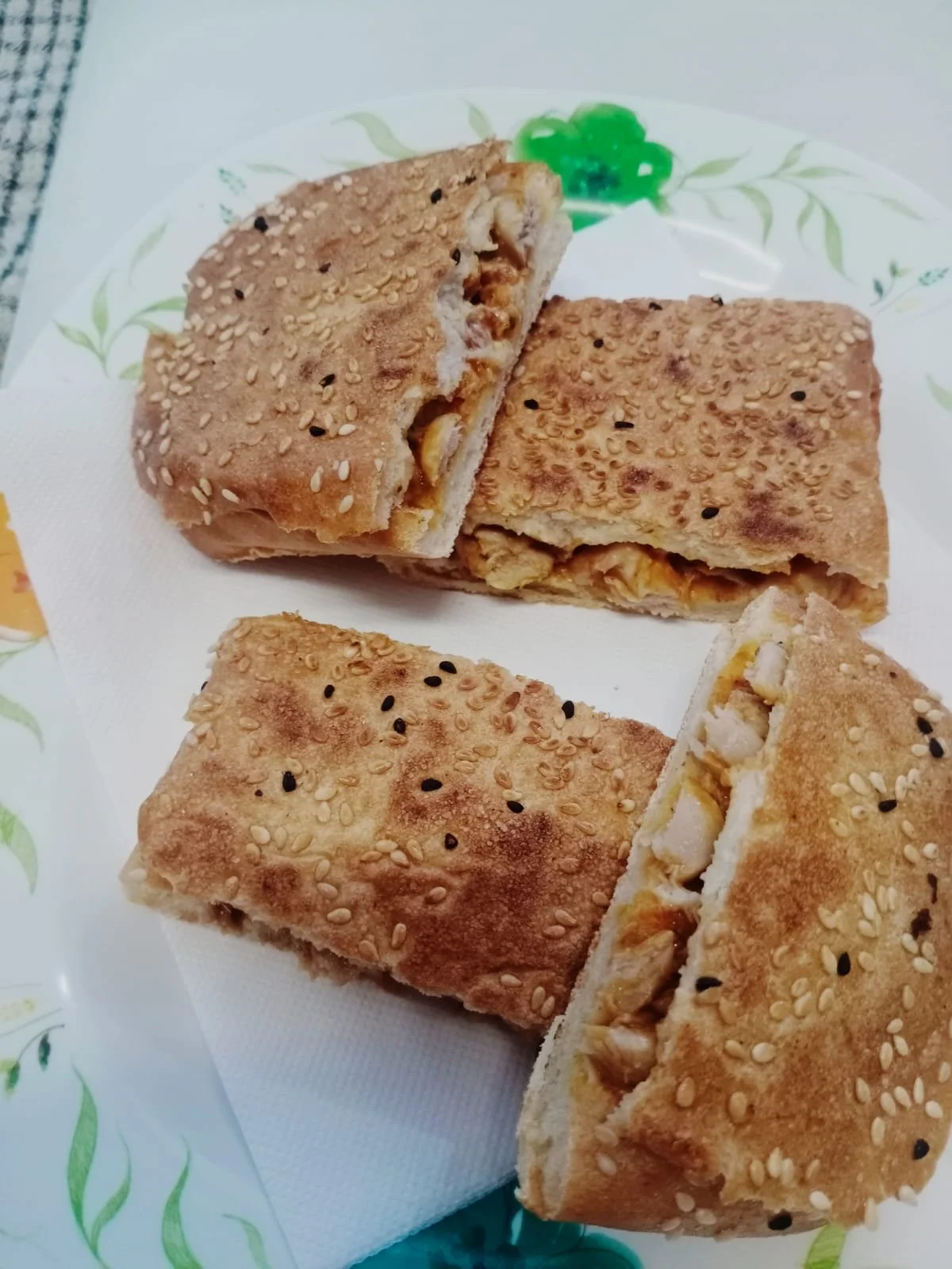 Three pieces of stuffed flatbread with sesame and black seeds on top served on a white paper napkin with a decorative plate underneath.