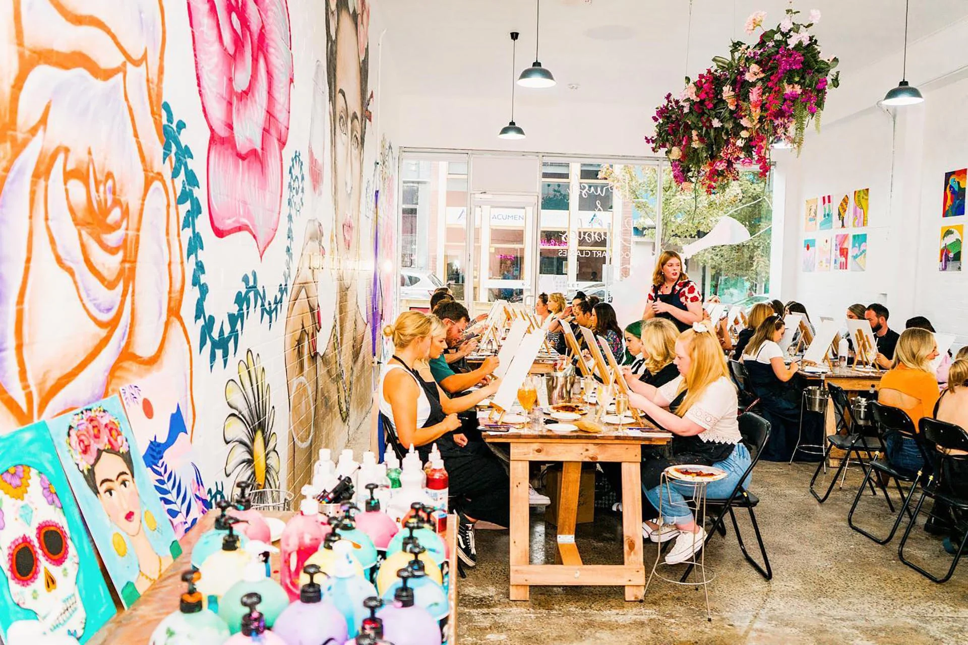 Women participating in a group painting activity in an art studio with colorful paintings on the walls, art supplies, and bottles of paint on tables.