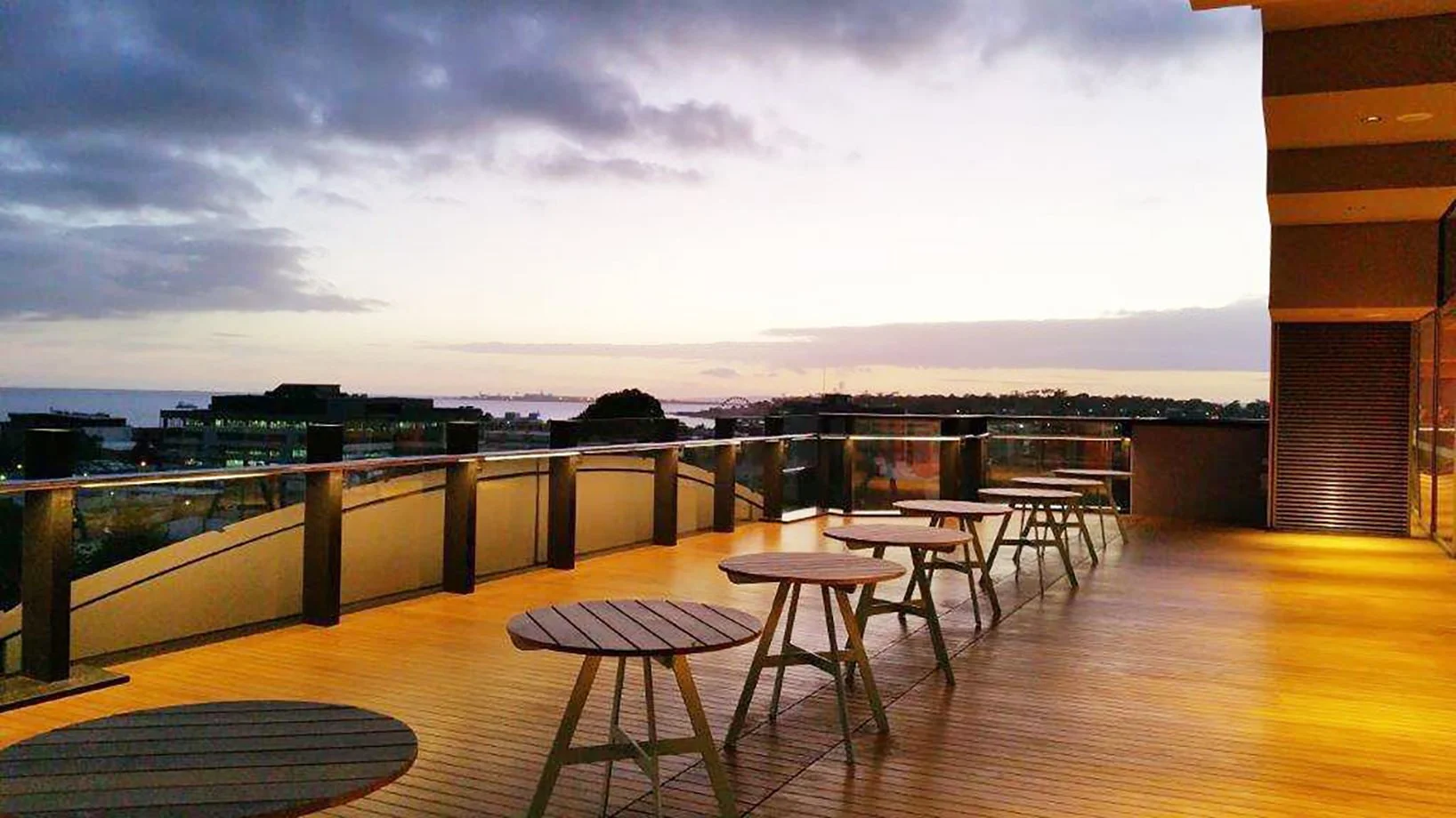 Empty outdoor balcony with wooden flooring, round tables, and stools, overlooking a city skyline at sunset with a cloudy sky.