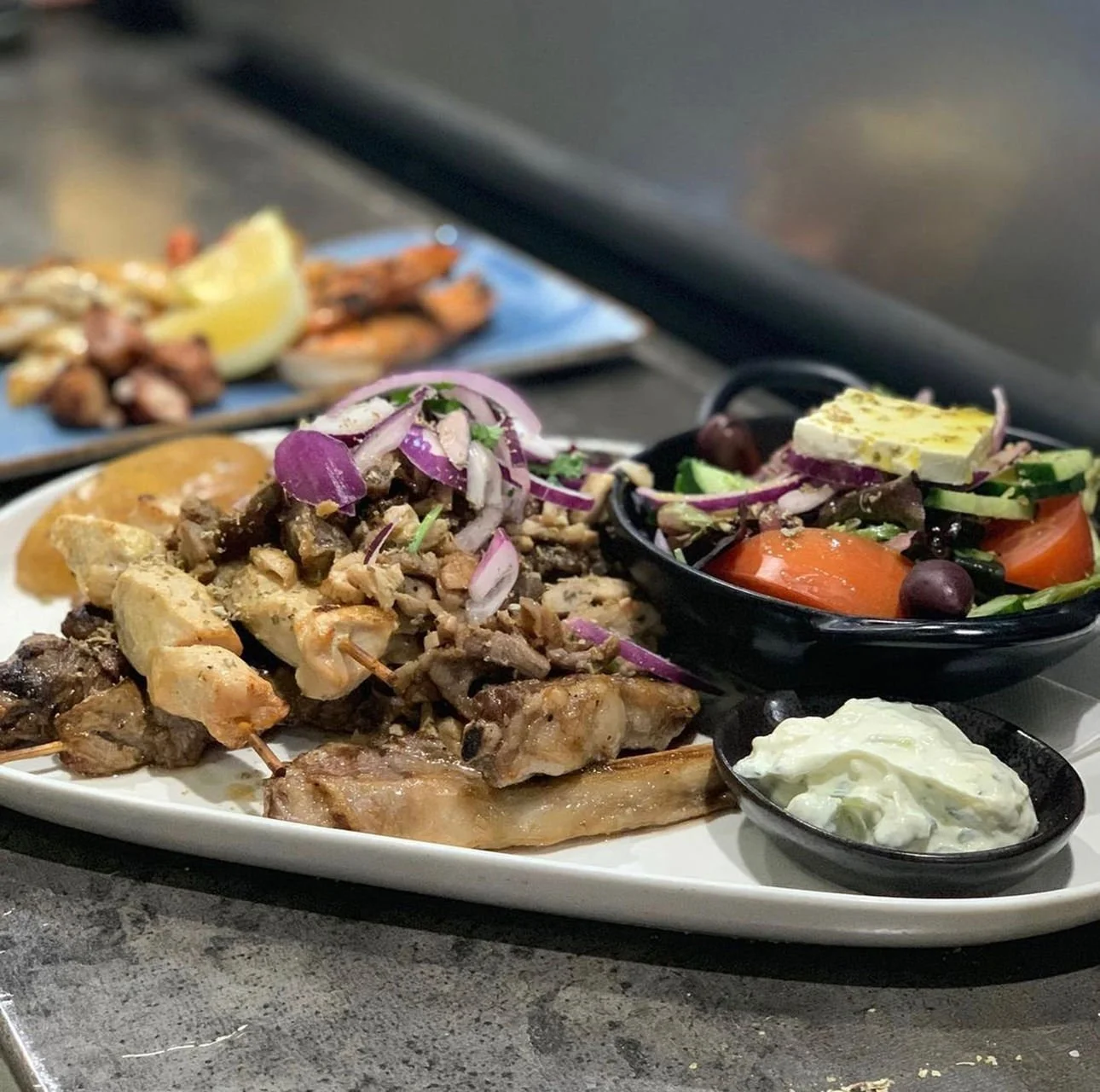 Plate with grilled meat, grilled vegetables, and side salads, including tomato, cucumber, olives, and feta cheese.