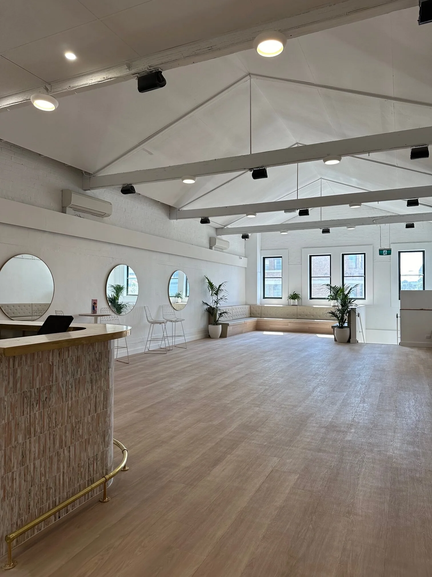 A spacious, well-lit room with a high vaulted ceiling, multiple circular mirrors on the wall, potted plants, and large windows letting in natural light. There are minimal furnishings including a wooden reception desk, a built-in seating area, and a f