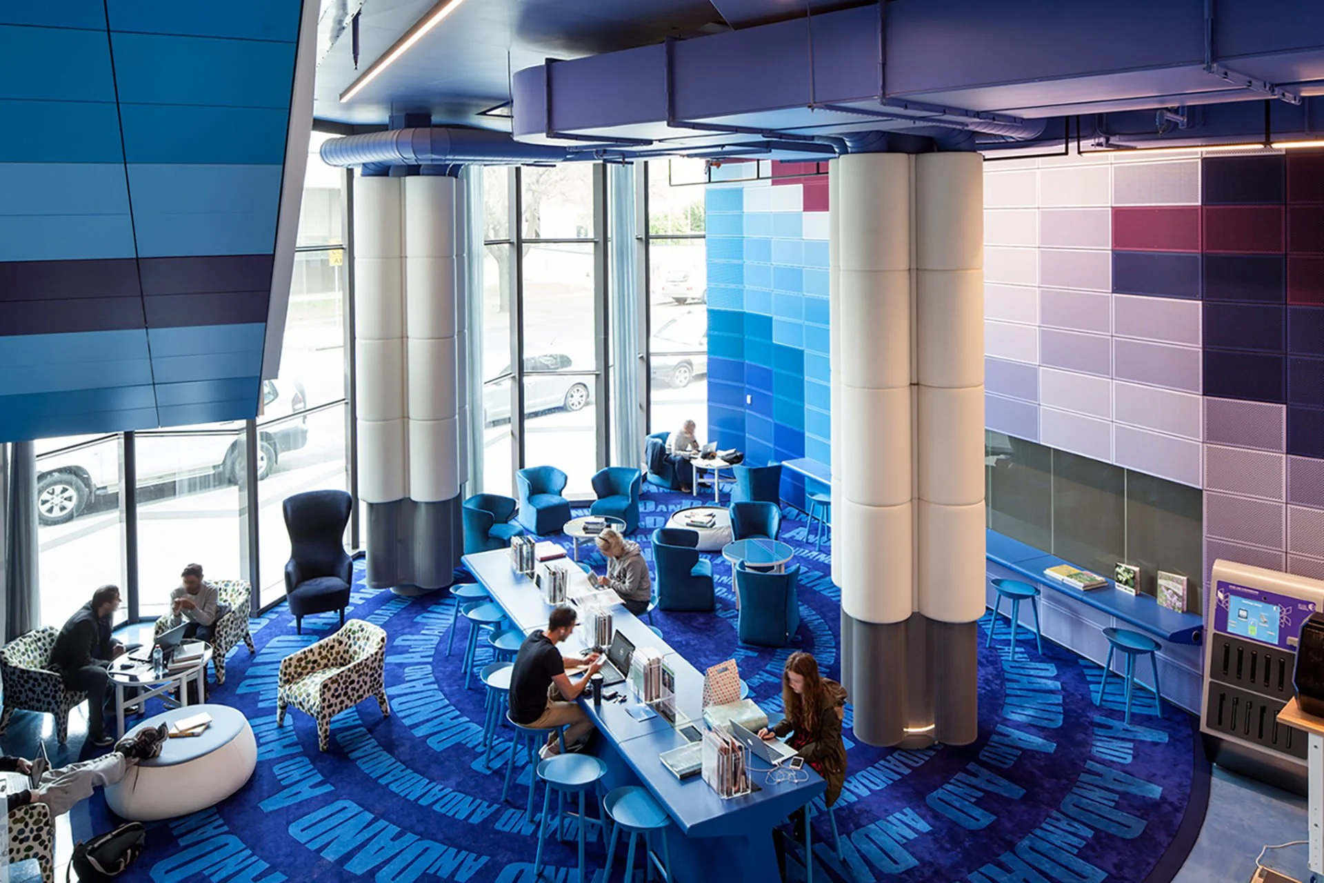 Modern lobby with large glass windows, blue and purple wall panels, and a blue patterned carpet. Several people are seated at tables and working on laptops or having conversations.