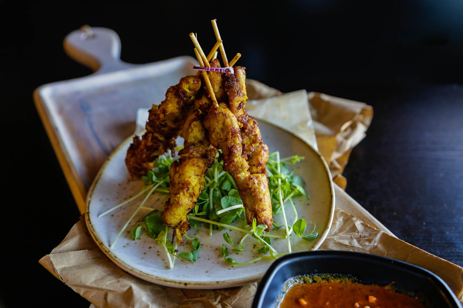 Fried chicken skewers on a bed of microgreens served on a ceramic plate, with a dipping sauce in a black dish nearby, placed on brown paper and a wooden tray.