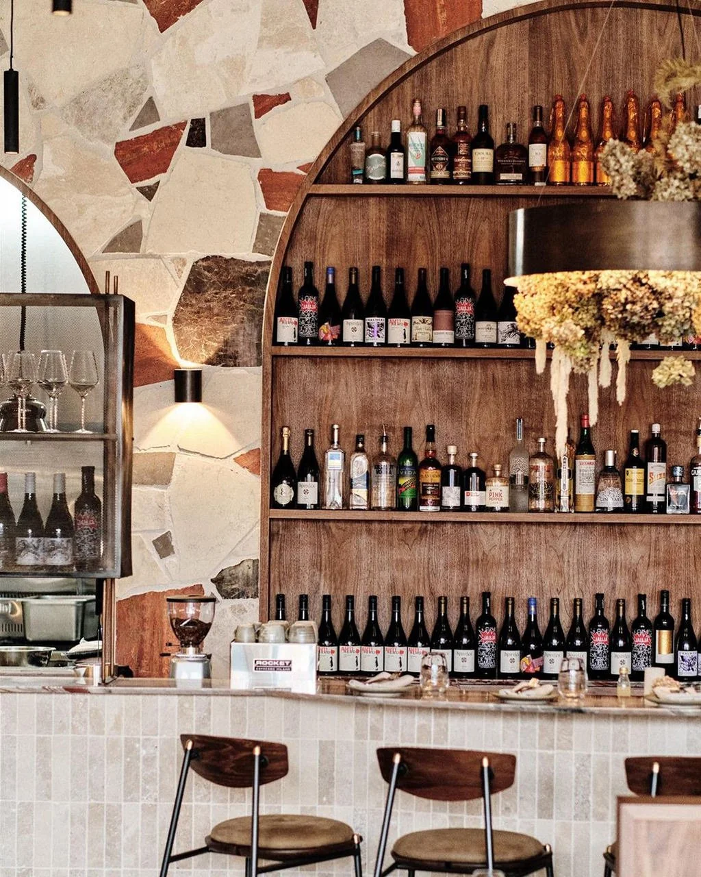 A wooden bar counter with shelves of wine bottles and liquor bottles, a stone wall behind it, and a hanging floral decoration. There are glasses and barware on the counter, with bar stools in front.