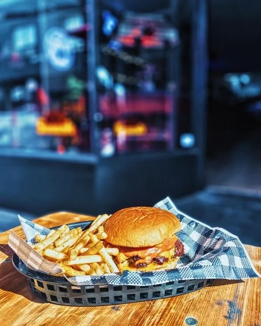 Burger with fries served in a black basket on a checkered paper liner on a wooden table, with a blurred storefront in the background.