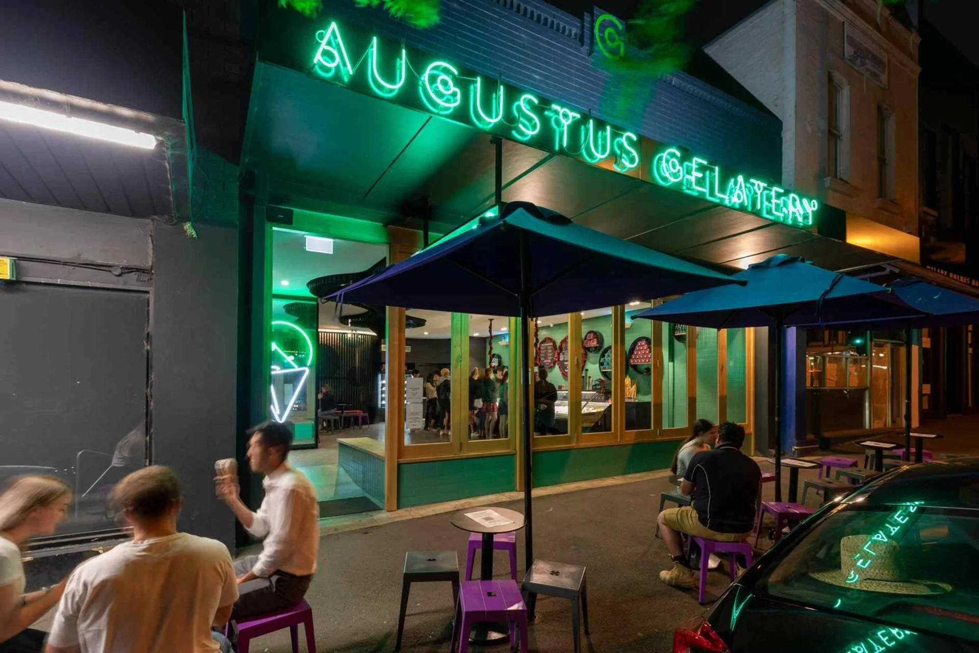 Night view of Augustus Gelato shop with neon sign, outdoor seating with purple stools, and people sitting outside.