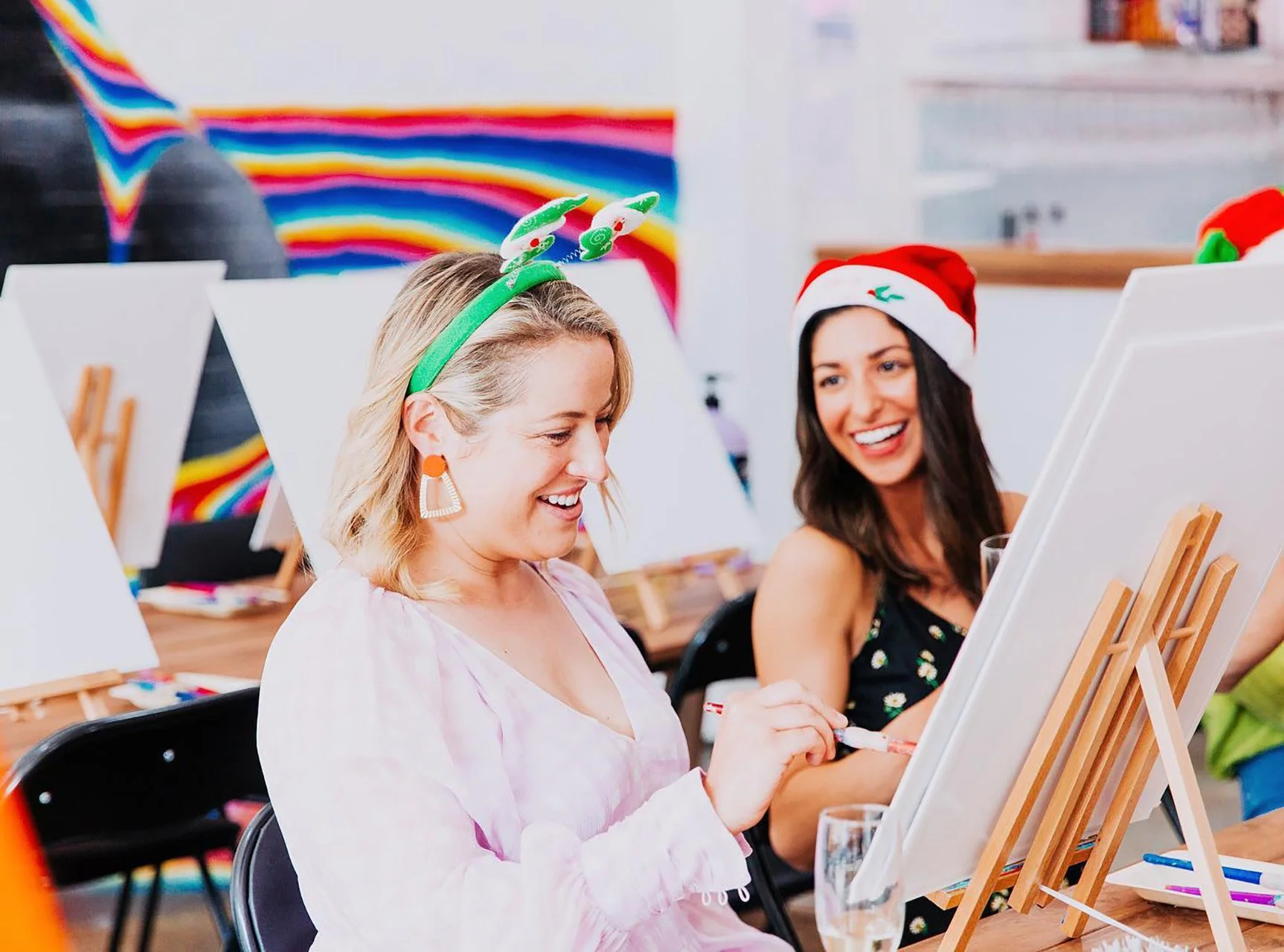 Two women celebrating Christmas at an art class, with holiday hats and festive headbands, painting on canvases set on easels, and colorful decorations in the background.