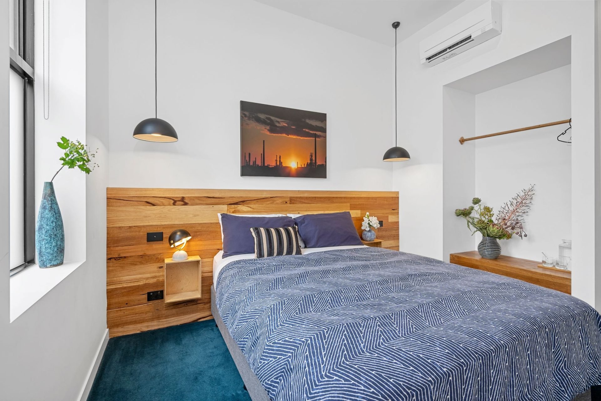 Modern bedroom with white walls, a wooden headboard, nautical-themed bedding, and decorative plants, featuring a wall-mounted TV and pendant lighting.