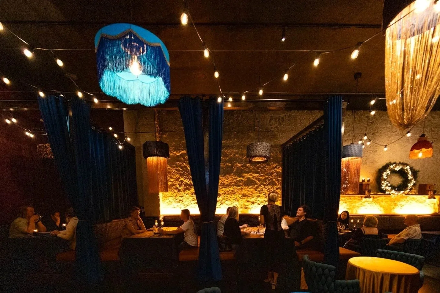 A dimly lit restaurant interior with black curtains, string lights, and a large teal chandelier. People are seated at tables, and there is a decorated wreath on the back wall. The decor is warm and inviting, suitable for an evening gathering or dinne
