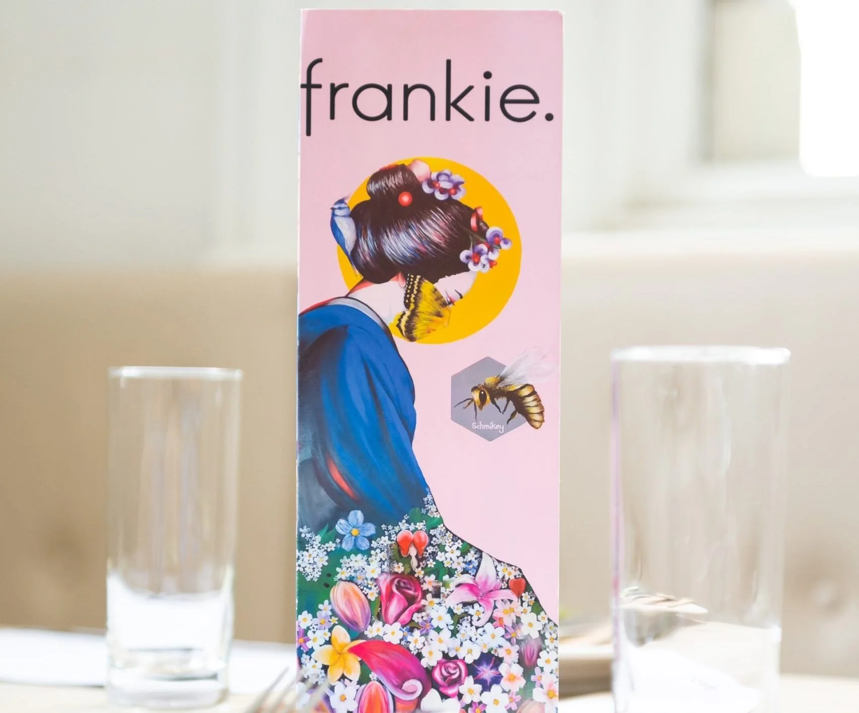 A pink vertical banner with the word 'frankie.' at the top, featuring a woman with a traditional Japanese hairstyle adorned with flowers, surrounded by a yellow circle. A butterfly and a bee are included in the design, along with flowers at the botto