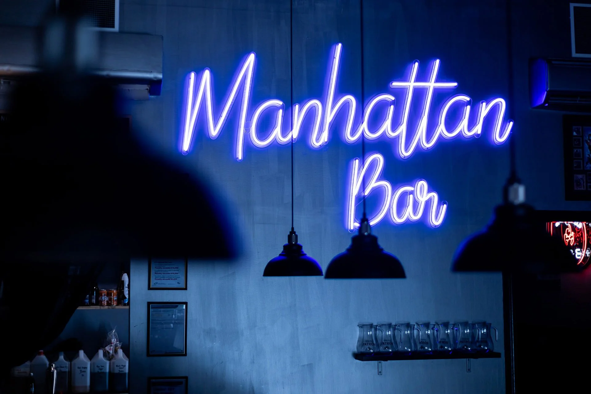Blue neon sign reading 'Manhattan Bar' on a gray wall inside a bar, with black pendant lights hanging below the sign.