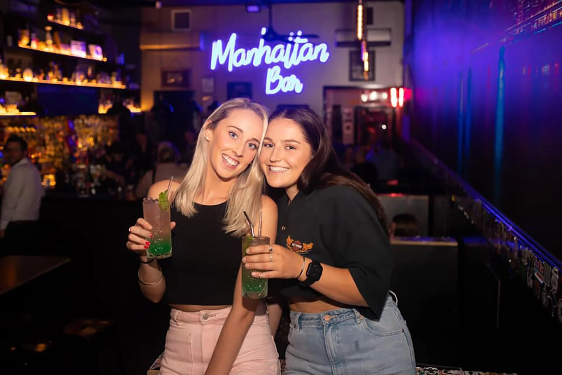 Two women smiling and holding colorful cocktails at a bar with neon sign that reads 'Marmahian Bar' in the background.