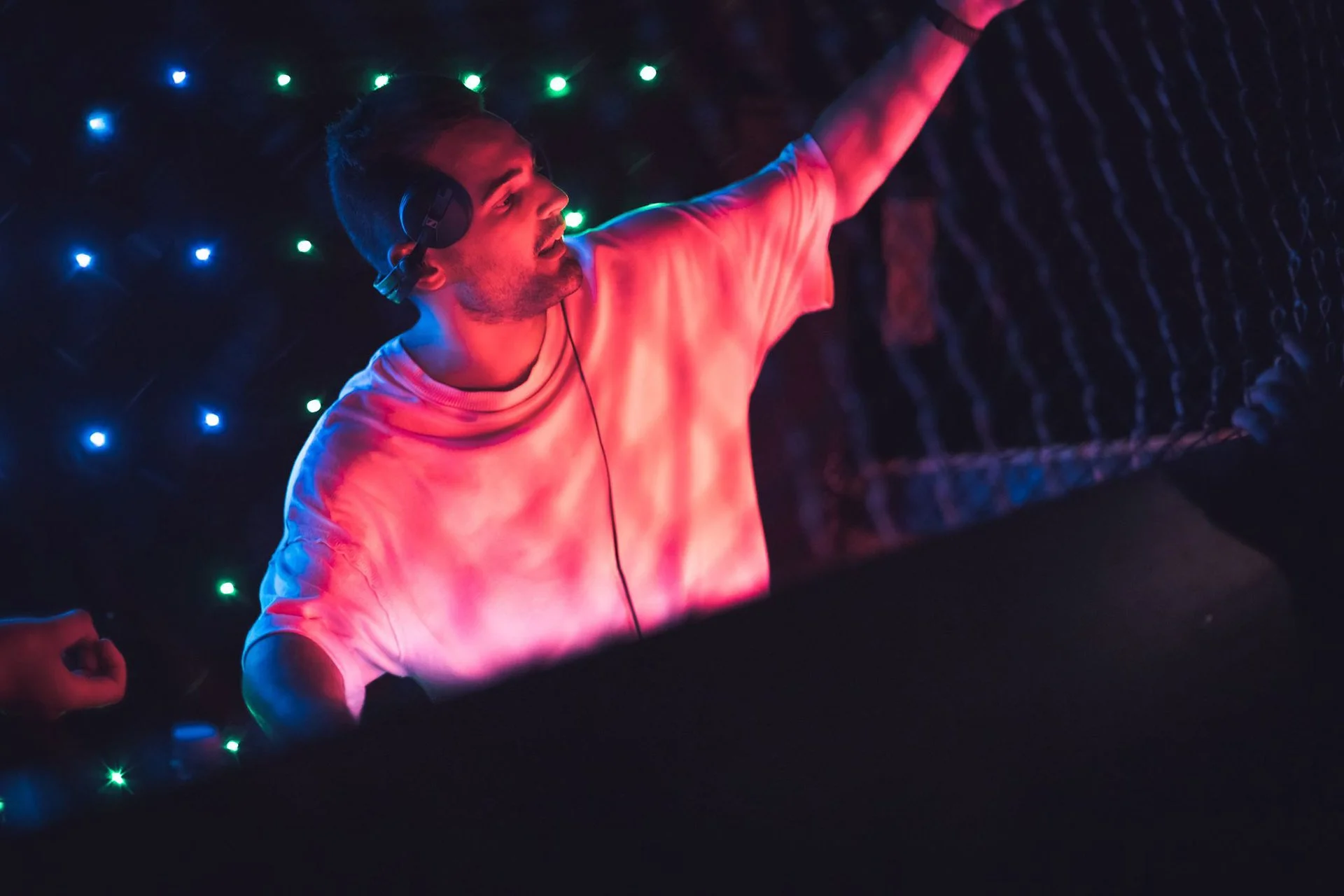 A DJ wearing headphones is performing at a nightclub with colorful lights in the background.